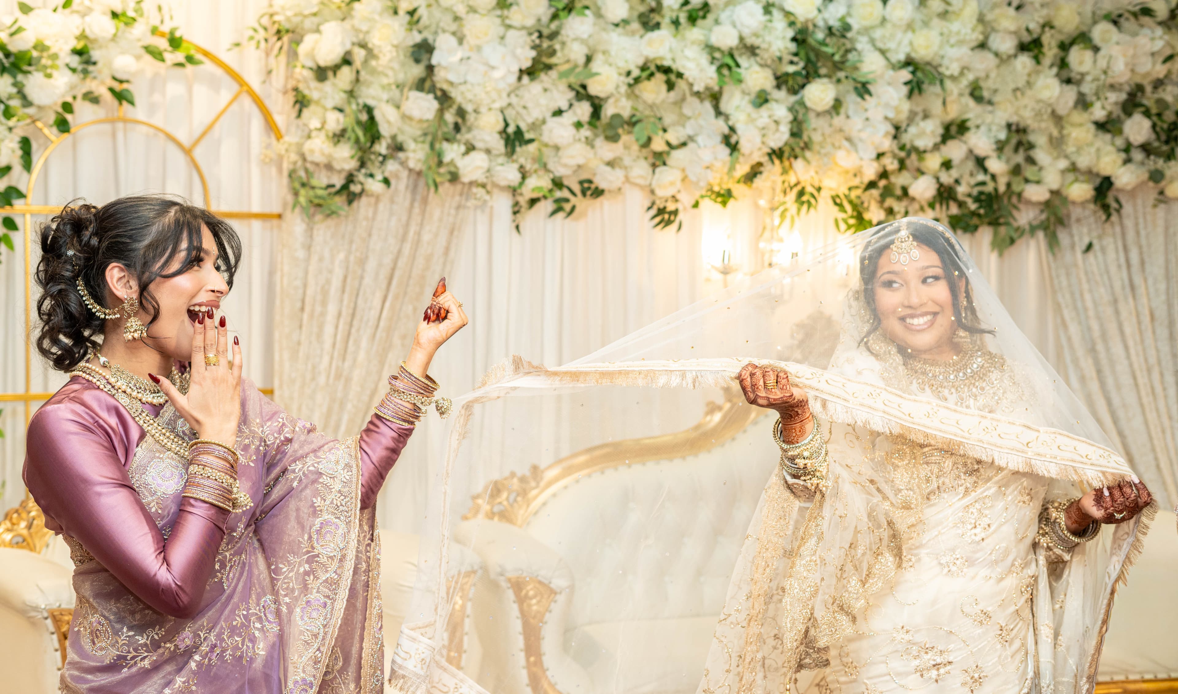 Two women smiling beneath a raised veil on a white-and-gold wedding stage.
