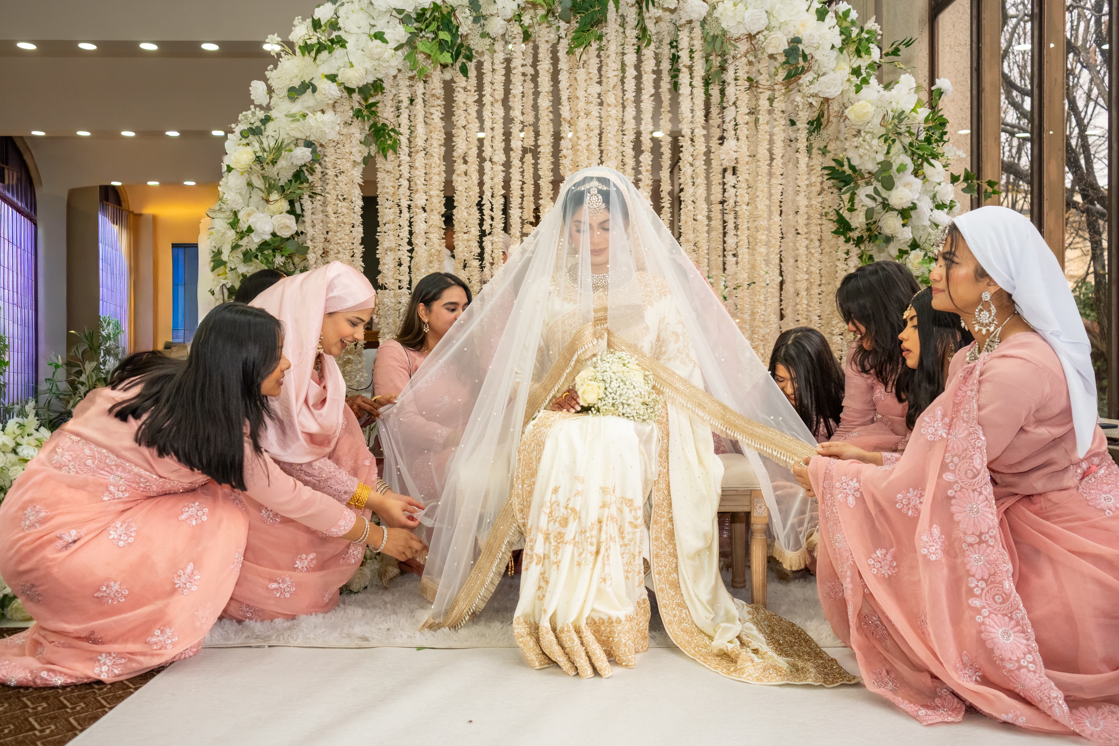 Bride seated beneath a floral installation while attendants arrange her veil.